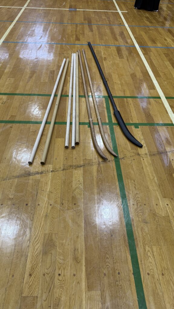 Wooden staffs, practice swords and a padded polearm arranged on a dojo floor.