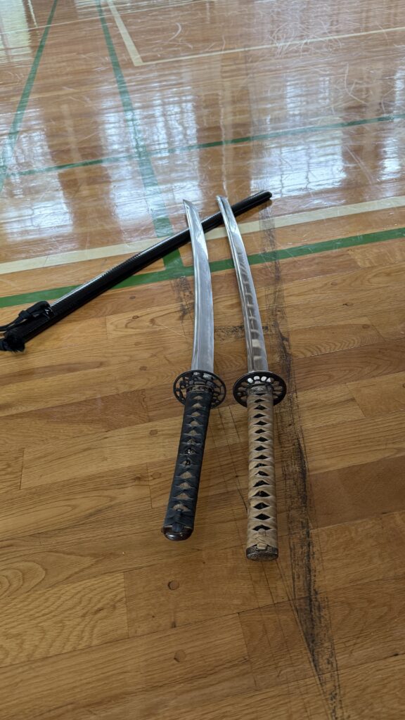 Two Japanese swords and a black scabbard on a wooden dojo floor.